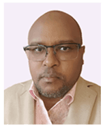 Bald man with glasses wearing a beige jacket and pink shirt looks directly forward.